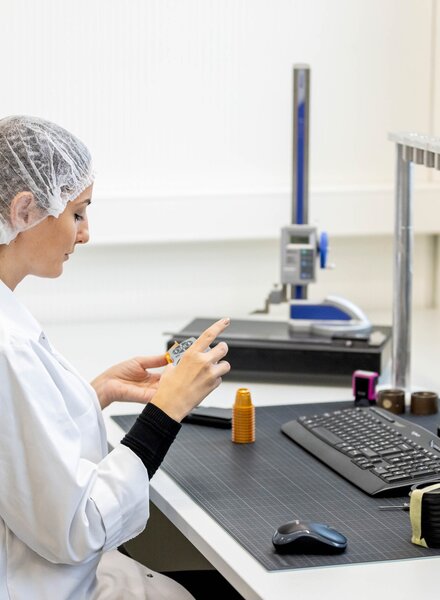 Mitarbeiterin im weissen Laborkittel und Haarnetz prüft Kunststoffkomponenten im Prüflabor von säntis packaging – Qualitätskontrolle und Bemusterung mit digitaler Dokumentation.