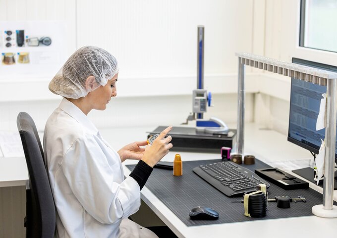 Mitarbeiterin im weissen Laborkittel und Haarnetz prüft Kunststoffkomponenten im Prüflabor von säntis packaging – Qualitätskontrolle und Bemusterung mit digitaler Dokumentation.