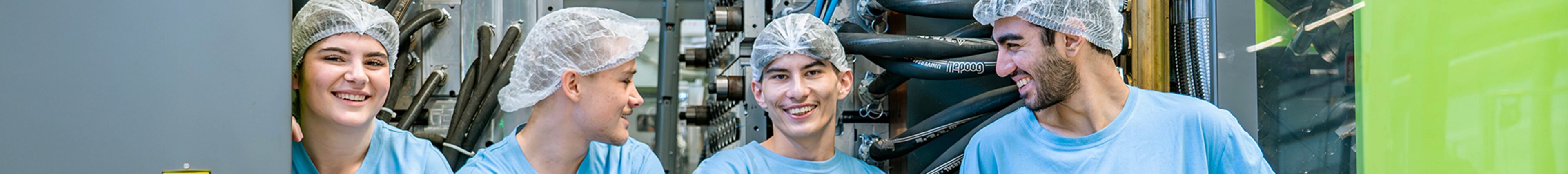 Vier Mitarbeitende von säntis packaging in blauen Arbeitskleidern lachen gemeinsam in der Produktionshalle – steht für Teamgeist, Freude an der Arbeit und eine offene Unternehmenskultur.