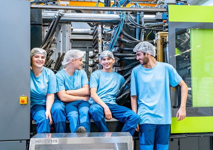 Vier Lernende in blauer Arbeitskleidung sitzen lachend vor einer modernen Spritzgussmaschine bei säntis packaging – Ausbildung in einem technologischen und teamorientierten Umfeld.