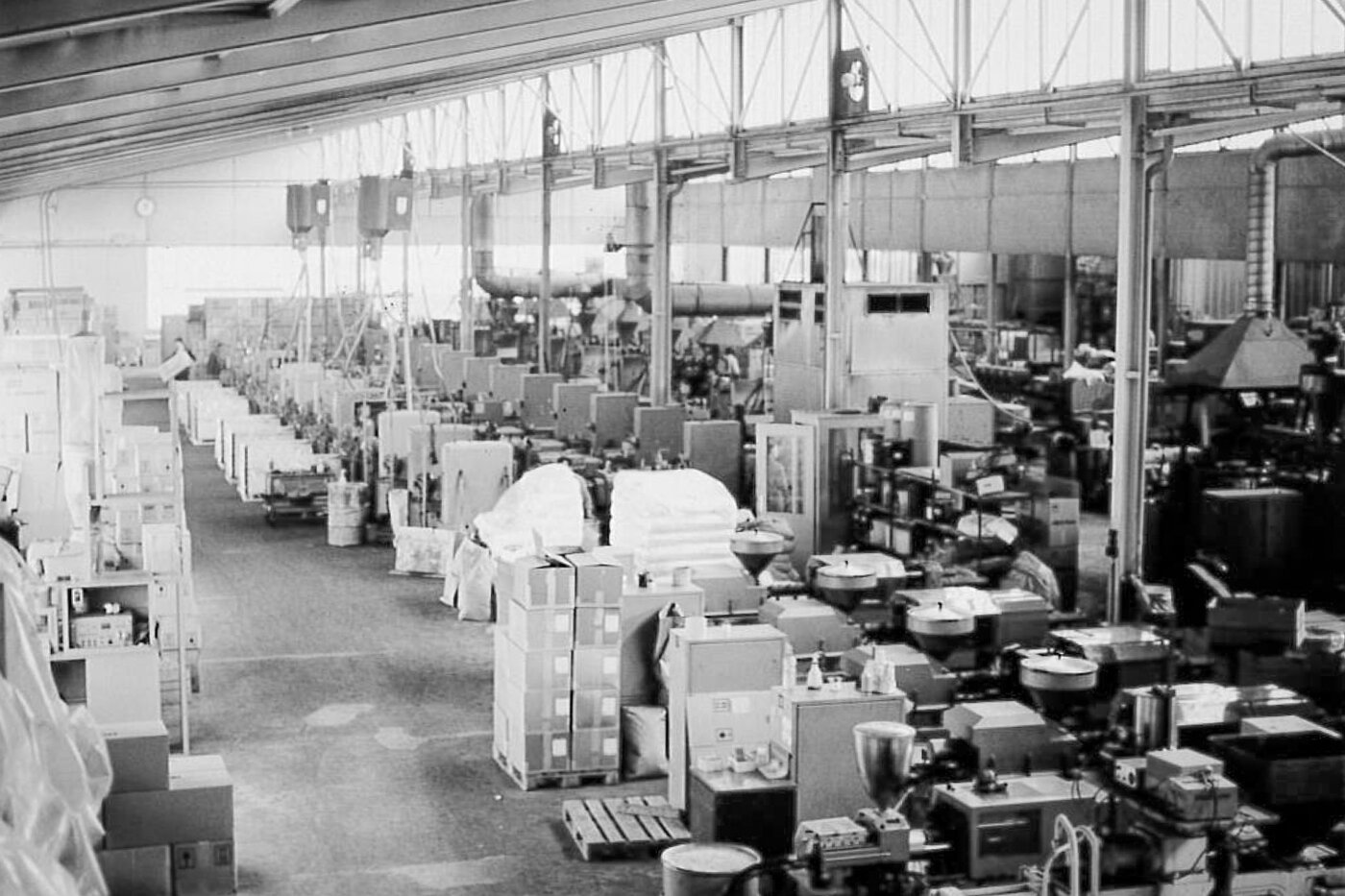 Schwarz-Weiss-Fotografie einer Produktionshalle aus den 1950er-Jahren: Reihen von Spritzgussmaschinen und Verpackungsmaterialien zeigen den Beginn der industriellen Herstellung von Joghurtbechern aus Polystyrol (PS) bei SÄNTIS – ein Meilenstein in der Schweizer Lebensmittelverpackung.