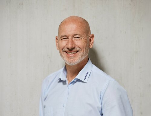 Porträt von Elmar Bösch, CFO & HR Director bei säntis packaging ag – in hellblauem Hemd mit Firmenlogo, freundlich lächelnd und professionell vor hellem Hintergrund fotografiert.