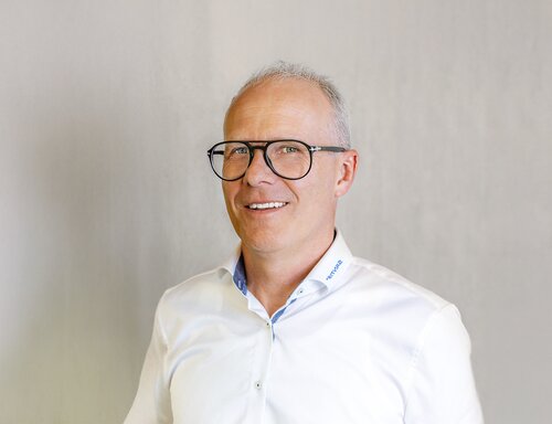 Porträt von Peter Amrein, Head of Business Development & R&D bei säntis packaging ag – in weissem Hemd mit Firmenlogo, mit Brille und offenem Lächeln vor hellem Hintergrund fotografiert.