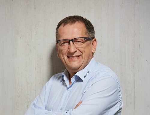 Porträt von Udo Kreissl, Head of Production bei säntis packaging ag – in hellblauem Hemd mit Firmenlogo, freundlich lächelnd und mit verschränkten Armen vor hellem Betonhintergrund fotografiert.