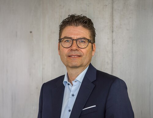 Porträt von Michael Thüler, CEO der säntis packaging ag – in dunkelblauem Sakko und hellblauem Hemd, mit Brille und professionellem Ausdruck vor hellem Hintergrund fotografiert.