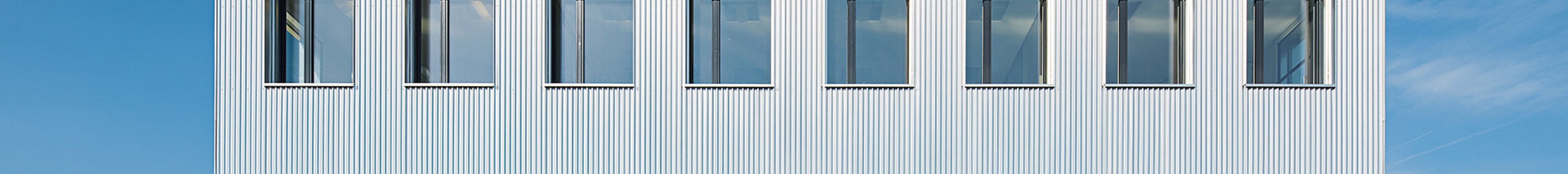 Moderne weisse Firmenfassade mit regelmässig angeordneten Fenstern unter blauem Himmel – Unternehmensgebäude von säntis packaging, Symbol für Struktur, Präzision und Transparenz.