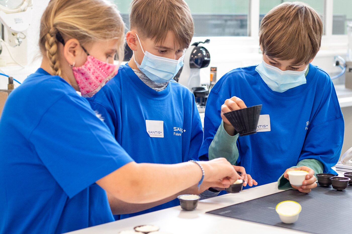 Drei Kinder in blauen Säntis Packaging T-Shirts messen Kunststoffteile während der Young-Tec-Days – praktische Einführung in Spritzguss und Qualitätskontrolle.