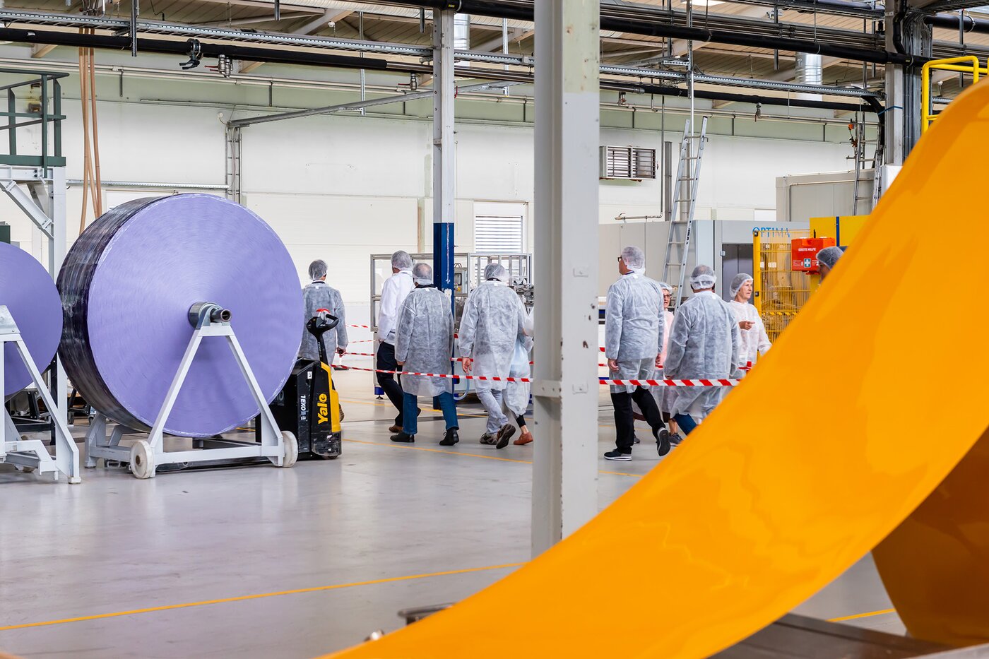 Besucherinnen und Besucher in Schutzkleidung bei einer Werksführung durch die Produktionshalle von Säntis Packaging – Blick auf Fertigungsanlagen und grosse Folienrollen während der Jubiläumsfeier.